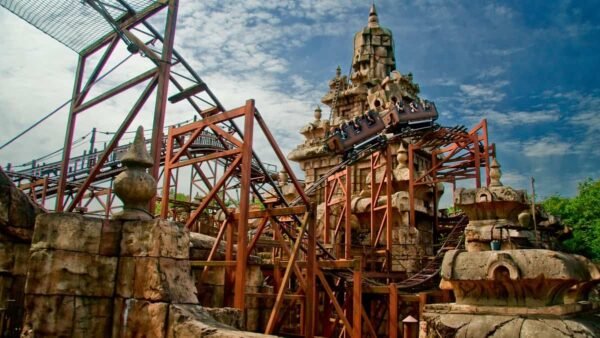 Une image d'une montagne russe avec un design de piste complexe, se faufilant autour et à travers une structure ornée en forme de tour ressemblant à des ruines antiques d'une aventure d'Indiana Jones, sur un ciel bleu avec des nuages.
