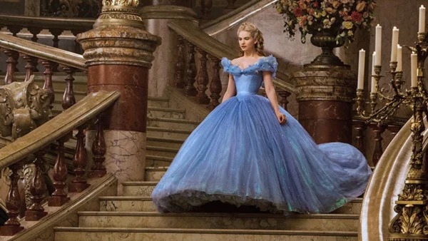 Une femme vêtue d'une élégante robe de bal bleue se tient sur un grand escalier dans une pièce luxueuse et ornée de bougies et d'un grand bouquet de fleurs, incarnant la grâce de Cendrillon.