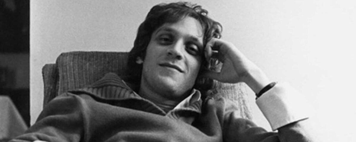 Photo en noir et blanc d'un jeune homme décontracté aux cheveux bouclés, légèrement souriant alors qu'il s'appuie sur un canapé, la main posée sur la tête. Il porte un pull décontracté Howard Ashman.