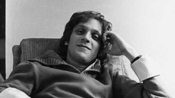 Photo en noir et blanc d'un jeune homme décontracté aux cheveux bouclés, légèrement souriant alors qu'il s'appuie sur un canapé, la main posée sur la tête. Il porte un pull décontracté Howard Ashman.