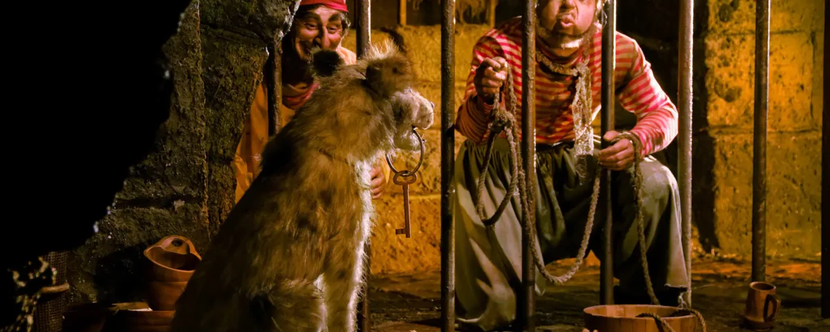 Dans un décor rustique et faiblement éclairé, deux artistes déguisés en pirates interagissent avec un gros chien assis entre eux. L’un tient une corde en souriant et l’autre regarde.