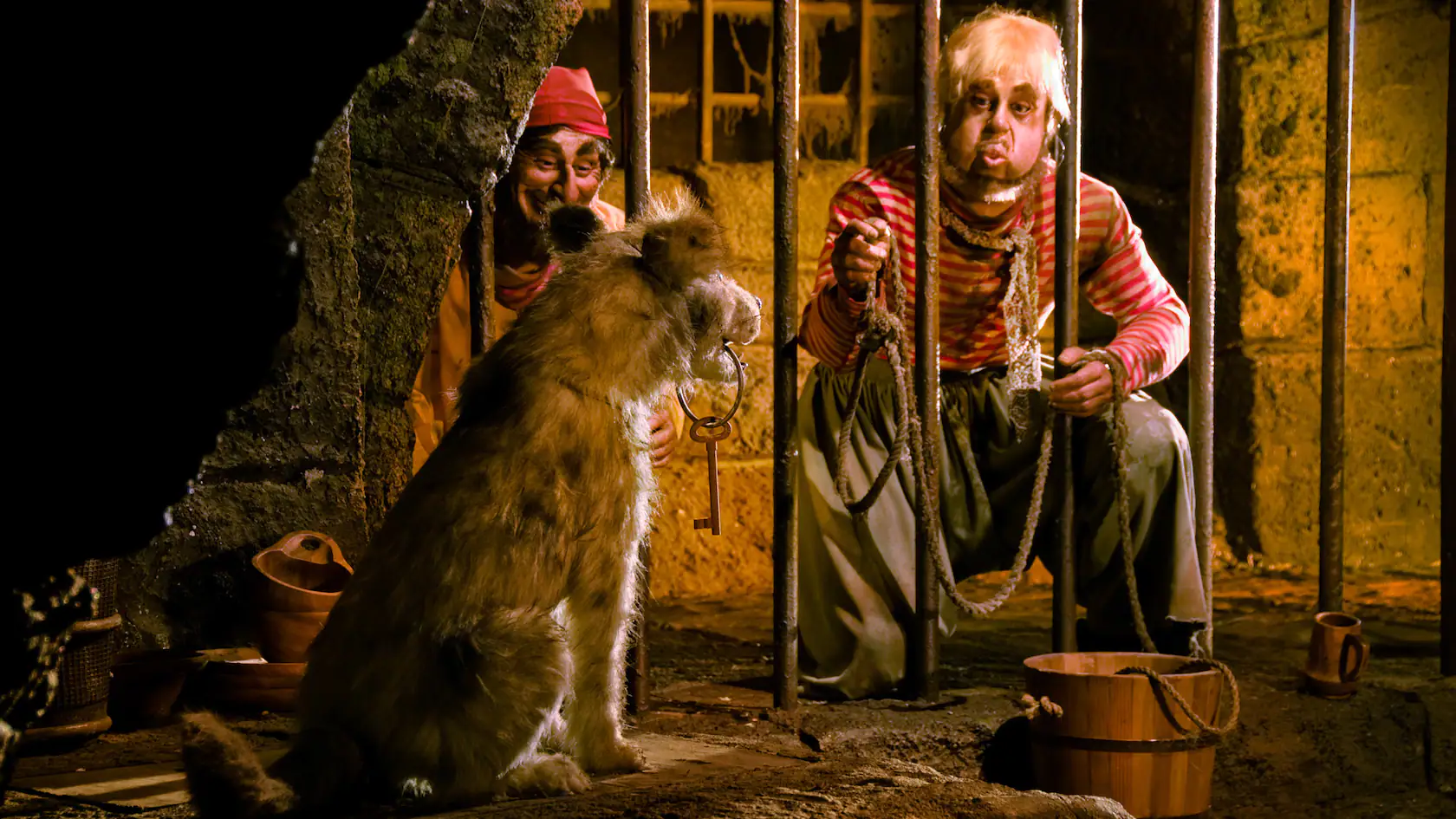 Dans un décor rustique et faiblement éclairé, deux artistes déguisés en pirates interagissent avec un gros chien assis entre eux. L’un tient une corde en souriant et l’autre regarde.