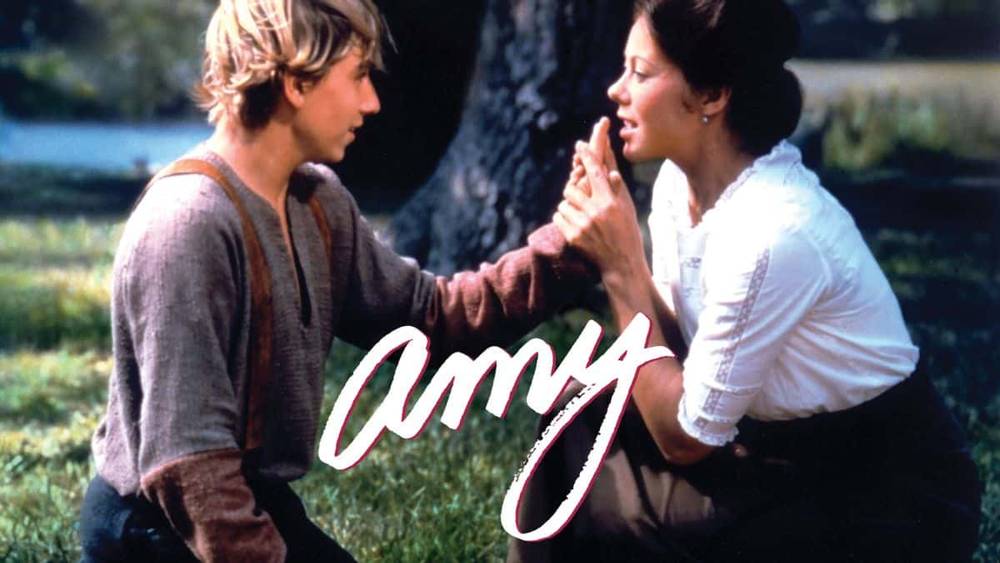 Un jeune homme et une femme se tiennent la main et se regardent sous un arbre. L'image a un éclairage doux et une sensation romantique, avec le mot « Amy » superposé en cursive rose.