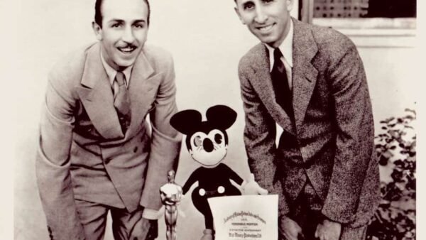 Deux hommes en costume souriant et posant avec une figurine Disney Mickey Mouse et une récompense sur une table entre eux, sur une photo vintage en noir et blanc.
