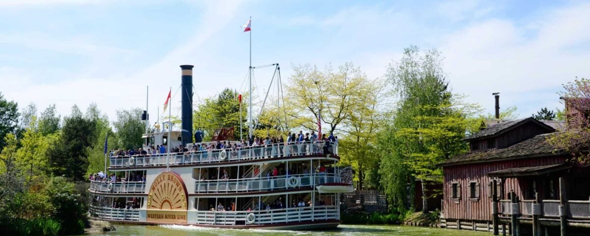 Un grand bateau à vapeur à aubes vintage rempli de passagers naviguant près d'un bâtiment en bois au bord d'une rivière à Disneyland par une journée ensoleillée avec une verdure luxuriante et un ciel bleu clair.