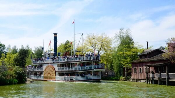 Un grand bateau à vapeur à aubes vintage rempli de passagers naviguant près d'un bâtiment en bois au bord d'une rivière à Disneyland par une journée ensoleillée avec une verdure luxuriante et un ciel bleu clair.