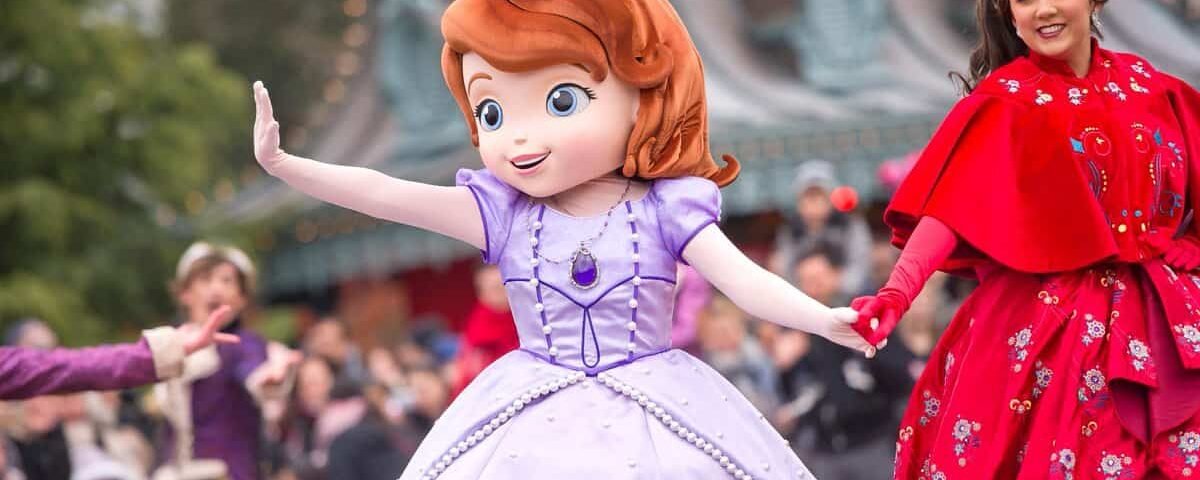 Une personne habillée en princesse Sofia salue la foule lors d'une "promenade princière", avec une actrice en tenue royale souriante derrière elle, dans un décor festif.