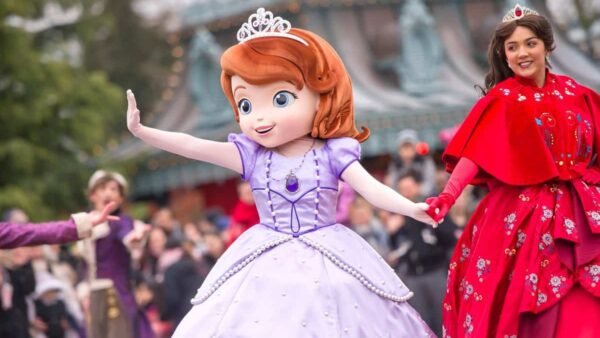 Une personne habillée en princesse Sofia salue la foule lors d'une "promenade princière", avec une actrice en tenue royale souriante derrière elle, dans un décor festif.