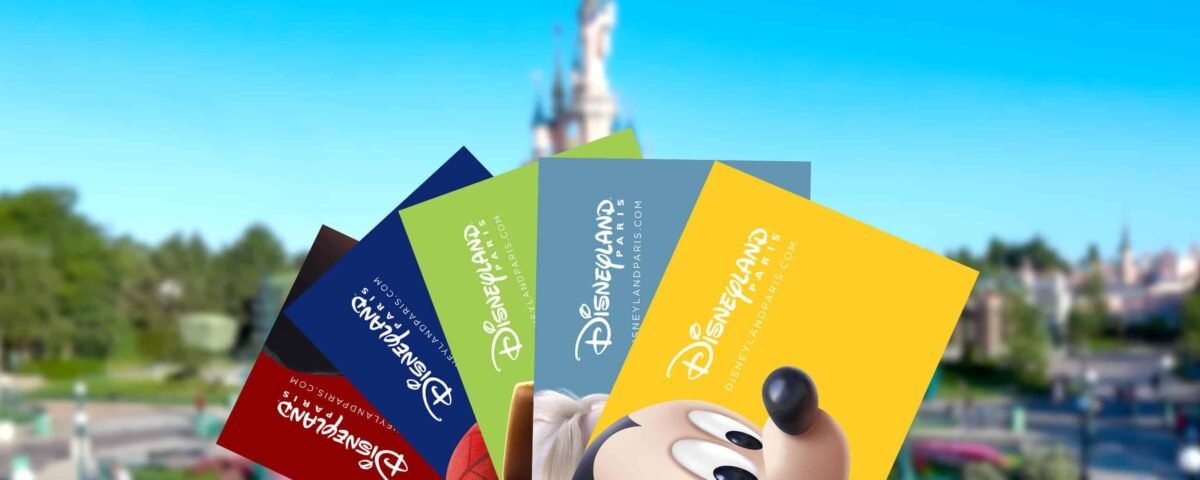 Une main tenant plusieurs billets colorés pour le parc Disneyland avec des tarifs ajustés, avec Mickey Mouse représenté sur l'un d'eux, sur un arrière-plan flou du parc Disneyland, y compris son château emblématique.