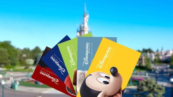 Une main tenant plusieurs billets colorés pour le parc Disneyland avec des tarifs ajustés, avec Mickey Mouse représenté sur l'un d'eux, sur un arrière-plan flou du parc Disneyland, y compris son château emblématique.