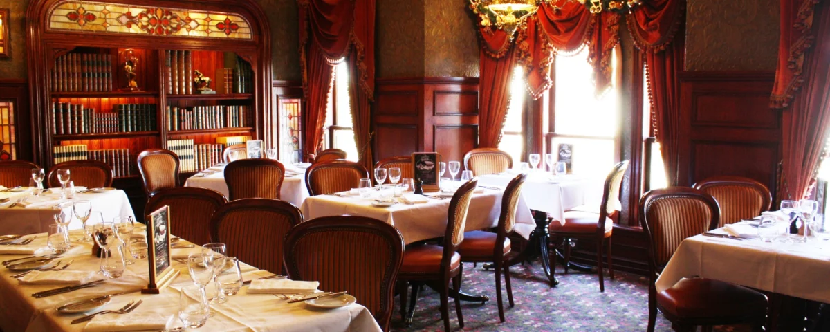 Salle à manger élégante avec tables dressées avec des draps blancs et de la vaisselle raffinée. Des bibliothèques en bois ornées, de riches draperies rouges et des lustres anciens rehaussent l'intérieur sophistiqué de style traditionnel.
