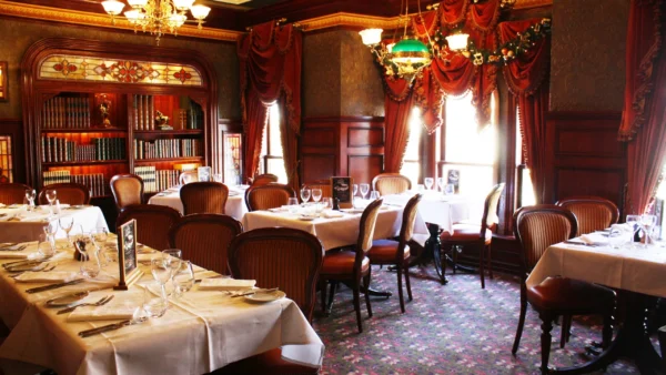 Salle à manger élégante avec tables dressées avec des draps blancs et de la vaisselle raffinée. Des bibliothèques en bois ornées, de riches draperies rouges et des lustres anciens rehaussent l'intérieur sophistiqué de style traditionnel.