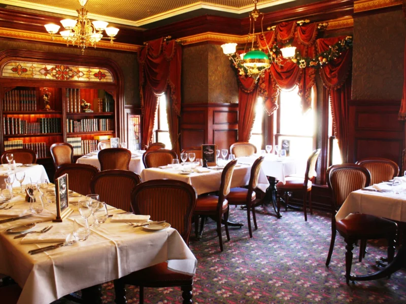 Salle à manger élégante avec tables dressées avec des draps blancs et de la vaisselle raffinée. Des bibliothèques en bois ornées, de riches draperies rouges et des lustres anciens rehaussent l'intérieur sophistiqué de style traditionnel.