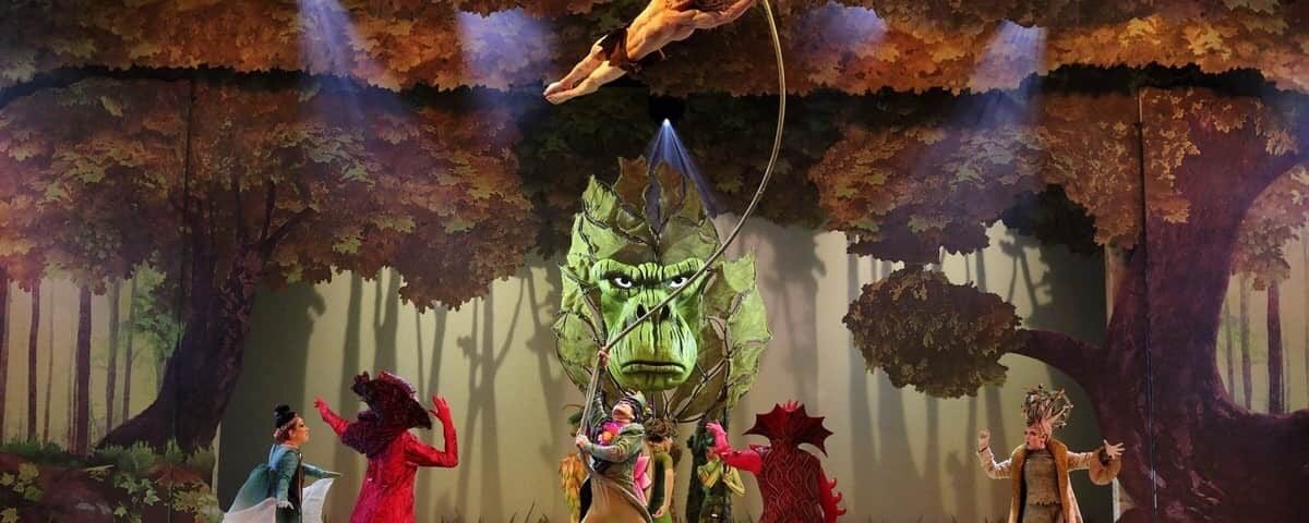Une scène théâtrale dans "La Forêt de l'Enchantement" avec des personnages aux costumes élaborés, dont un grand dragon vert et des interprètes habillés en arbres, au milieu des feuilles automnales.