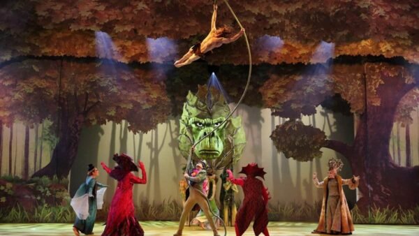 Une scène théâtrale dans "La Forêt de l'Enchantement" avec des personnages aux costumes élaborés, dont un grand dragon vert et des interprètes habillés en arbres, au milieu des feuilles automnales.