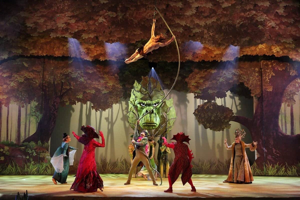 Une scène théâtrale dans "La Forêt de l'Enchantement" avec des personnages aux costumes élaborés, dont un grand dragon vert et des interprètes habillés en arbres, au milieu des feuilles automnales.