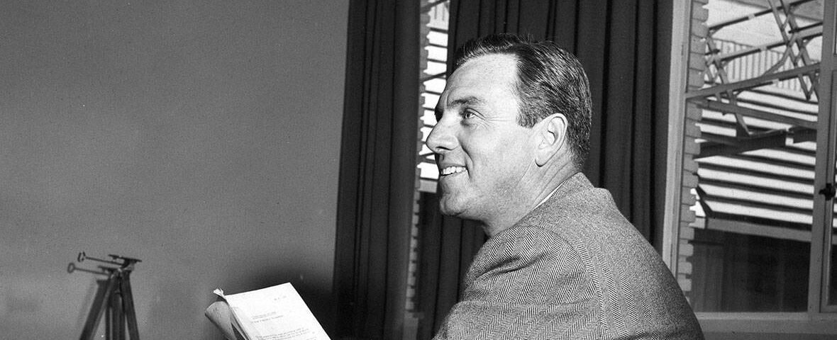 Photo en noir et blanc d'un homme souriant en costume, assis à un bureau, regardant sur le côté tout en tenant un script. Une fenêtre ouverte avec des stores est en arrière-plan. C'est David
