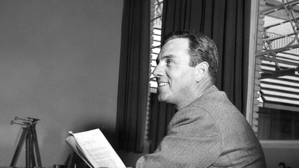 Photo en noir et blanc d'un homme souriant en costume, assis à un bureau, regardant sur le côté tout en tenant un script. Une fenêtre ouverte avec des stores est en arrière-plan. C'est David