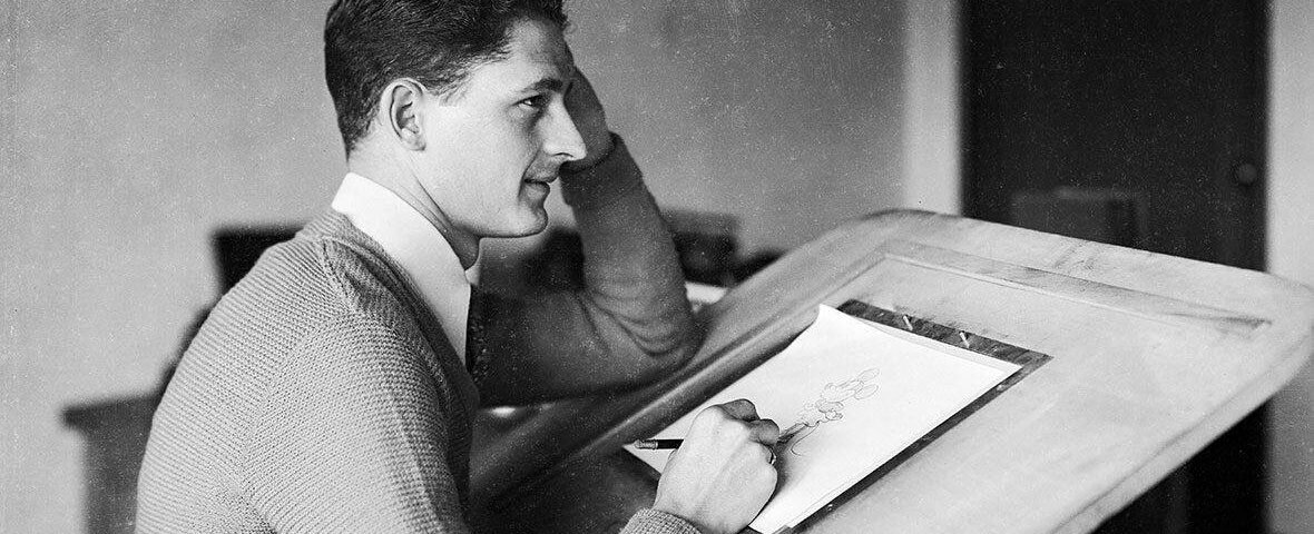 Une photo en noir et blanc de Les Clark, un jeune homme en pull, assis à une table à dessin et dessinant sur papier, posant son menton sur sa main, absorbé par son travail