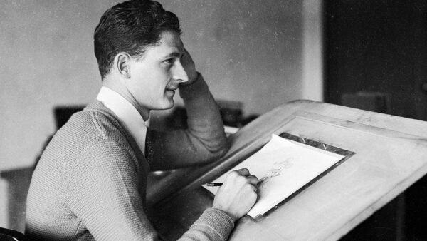 Une photo en noir et blanc de Les Clark, un jeune homme en pull, assis à une table à dessin et dessinant sur papier, posant son menton sur sa main, absorbé par son travail