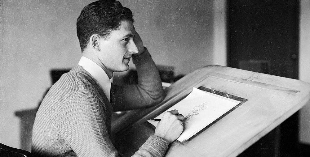 Une photo en noir et blanc de Les Clark, un jeune homme en pull, assis à une table à dessin et dessinant sur papier, posant son menton sur sa main, absorbé par son travail