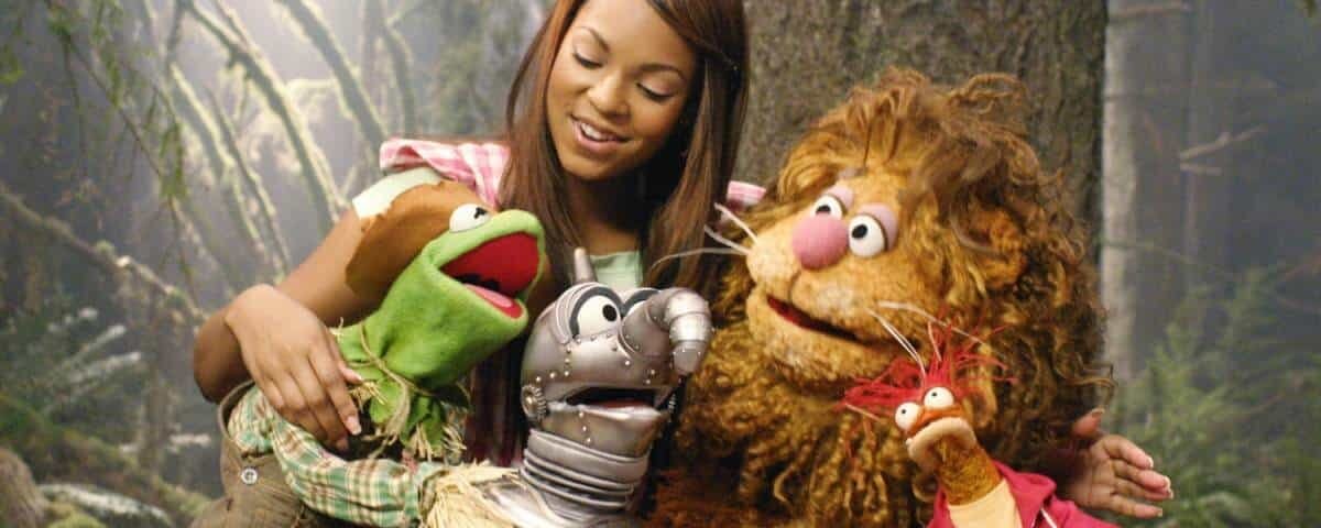 Une femme sourit en posant avec quatre personnages Muppet, dont Kermit la grenouille, dans un décor sur le thème de la forêt. Les marionnettes représentent différents animaux, ajoutant une atmosphère ludique.