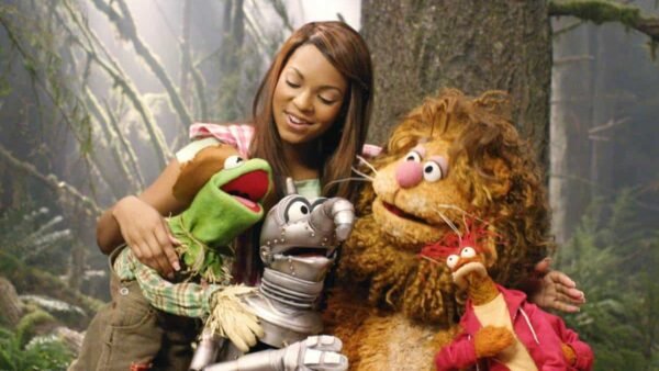 Une femme sourit en posant avec quatre personnages Muppet, dont Kermit la grenouille, dans un décor sur le thème de la forêt. Les marionnettes représentent différents animaux, ajoutant une atmosphère ludique.