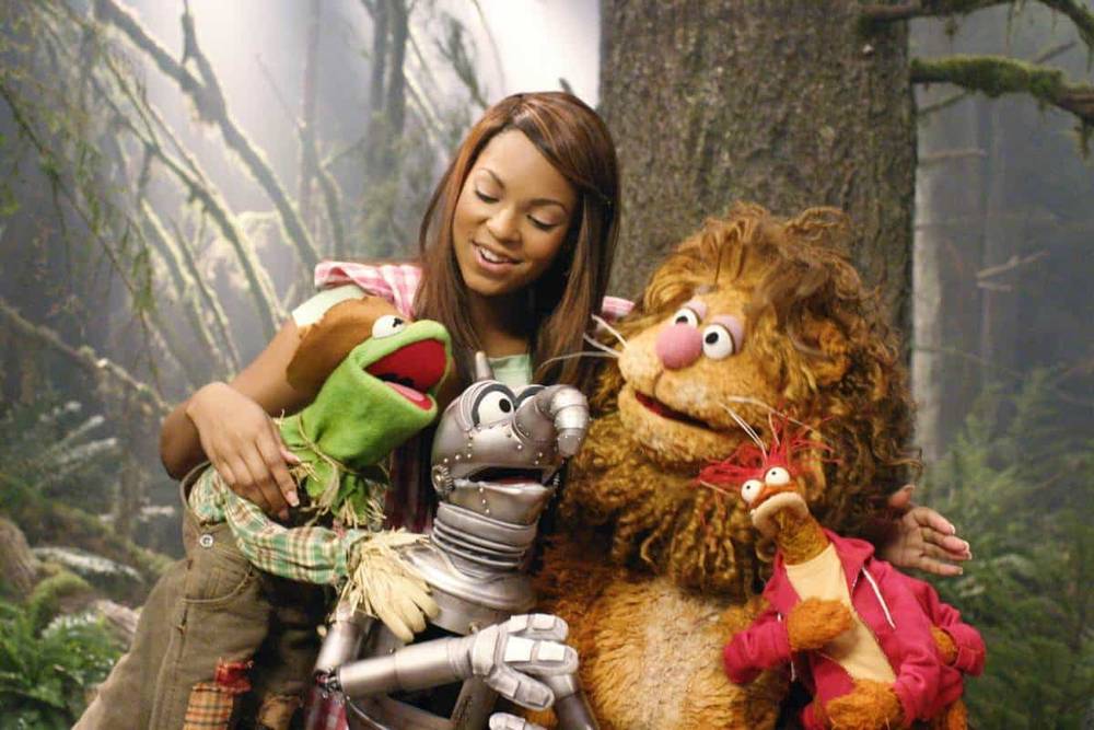 Une femme sourit en posant avec quatre personnages Muppet, dont Kermit la grenouille, dans un décor sur le thème de la forêt. Les marionnettes représentent différents animaux, ajoutant une atmosphère ludique.