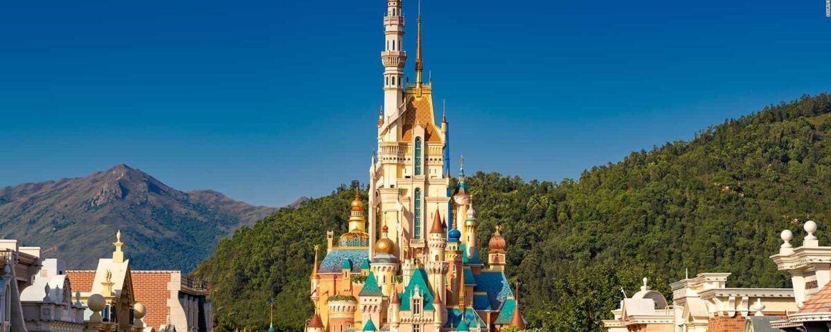 L'image montre le château vibrant et coloré de la Belle au bois dormant à Hong Kong Disneyland. C'est une journée ensoleillée et le majestueux château de conte de fées se dresse sur fond de ciel bleu clair et