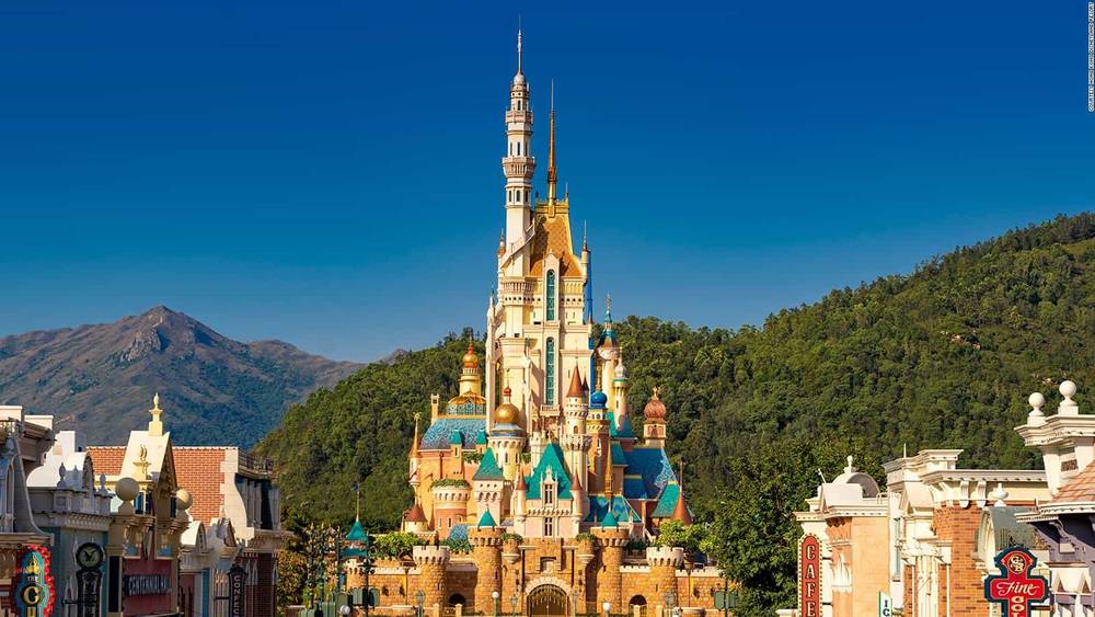 L'image montre le château vibrant et coloré de la Belle au bois dormant à Hong Kong Disneyland. C'est une journée ensoleillée et le majestueux château de conte de fées se dresse sur fond de ciel bleu clair et