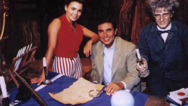 Trois individus (une femme, un homme en costume et un autre homme en blouse de laboratoire) se rassemblent autour d'une table avec une carte, une loupe et un globe dans un décor rempli d'accessoires sur le thème du défilé Disney.