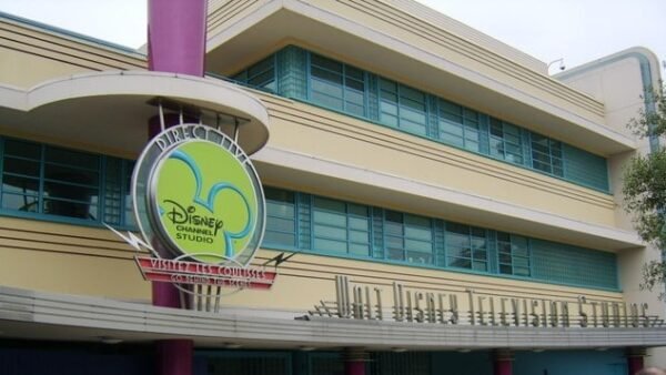 L'image montre l'entrée des studios de télévision Walt Disney, avec un logo circulaire vert et bleu, avec un panneau indiquant « rendez-nous visite sur www.disneychannel.com ». Le bâtiment a un rétro