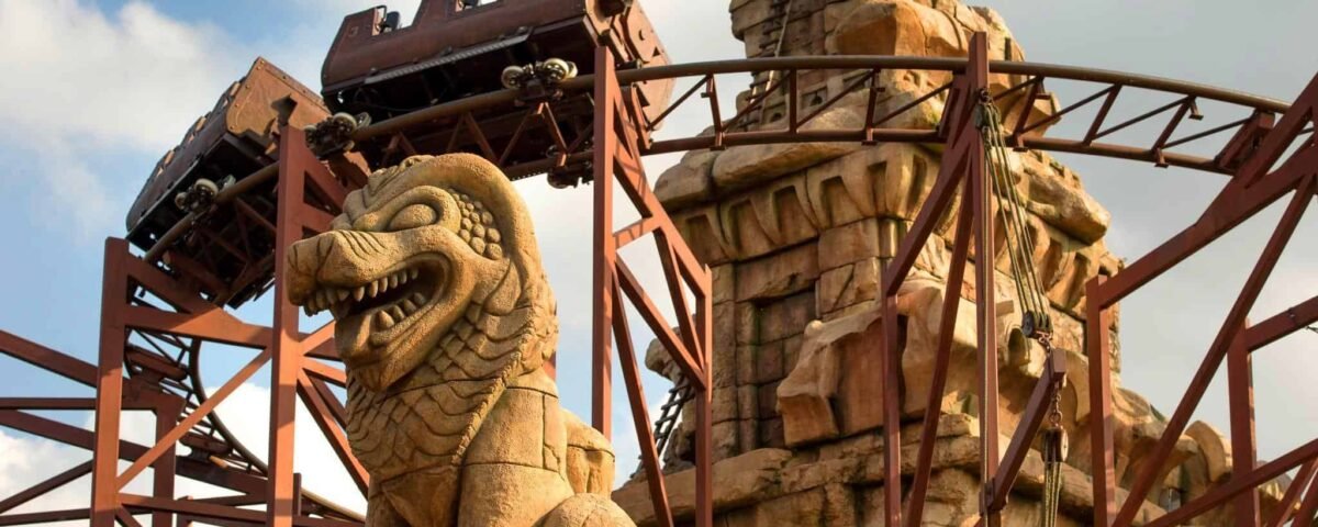 Un chariot de montagnes russes descendant une piste escarpée, encadré par de grandes structures ornementales en pierre, dont une statue proéminente d'une créature mythique ressemblant à un dragon dans "Indiana Jones et le Temple du Péril".