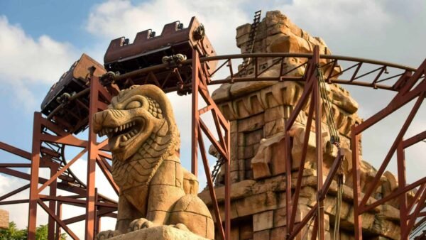 Un chariot de montagnes russes descendant une piste escarpée, encadré par de grandes structures ornementales en pierre, dont une statue proéminente d'une créature mythique ressemblant à un dragon dans "Indiana Jones et le Temple du Péril".