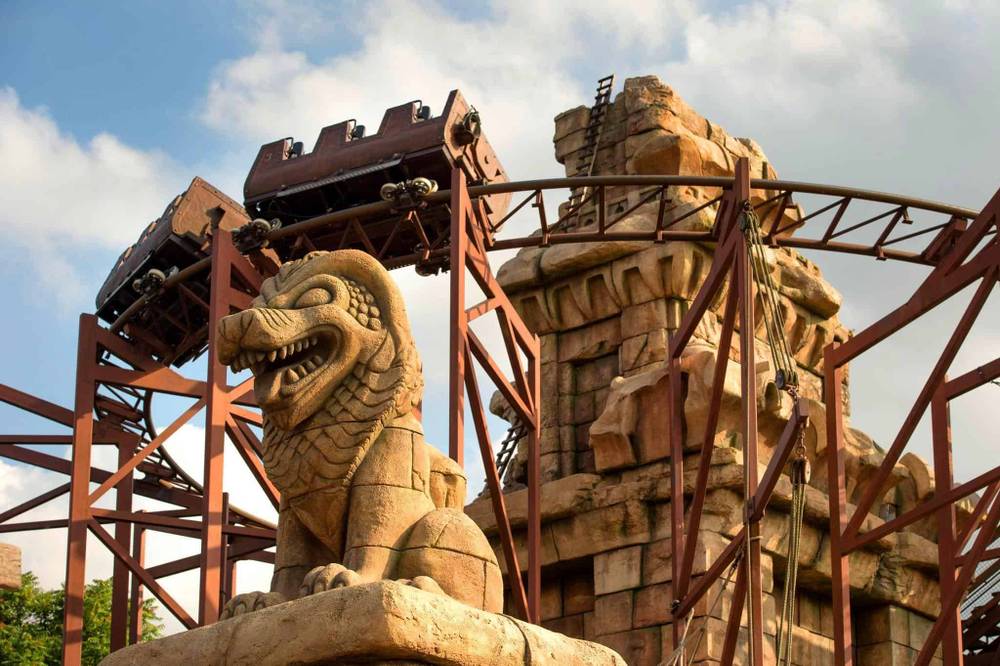 Un chariot de montagnes russes descendant une piste escarpée, encadré par de grandes structures ornementales en pierre, dont une statue proéminente d'une créature mythique ressemblant à un dragon dans "Indiana Jones et le Temple du Péril".