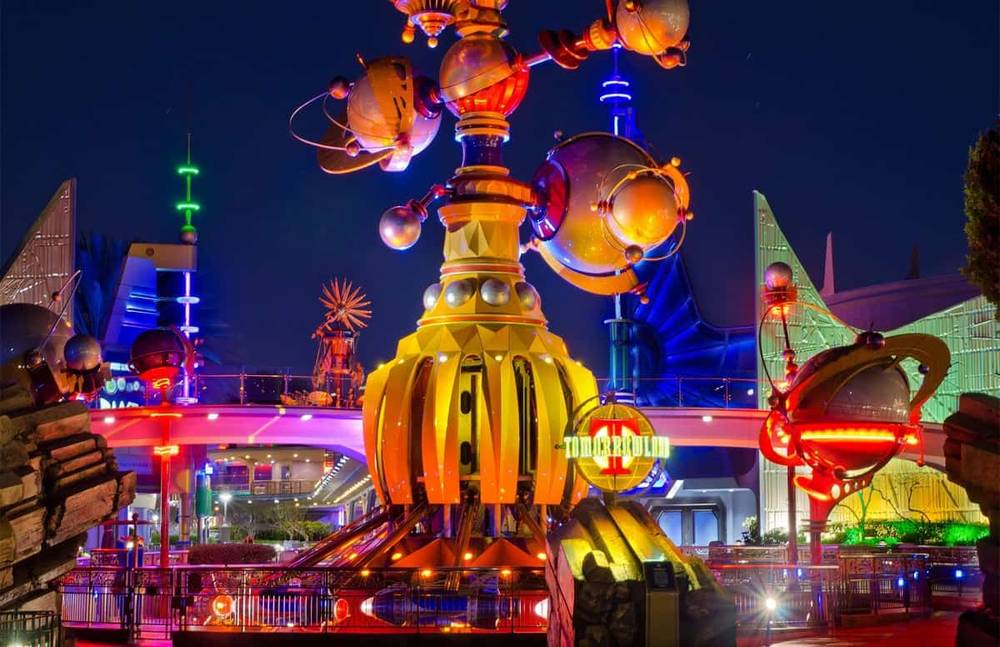Une scène nocturne vibrante de Tomorrowland à Disneyland, avec des bâtiments futuristes avec des lumières brillantes et une structure dorée proéminente au premier plan.
