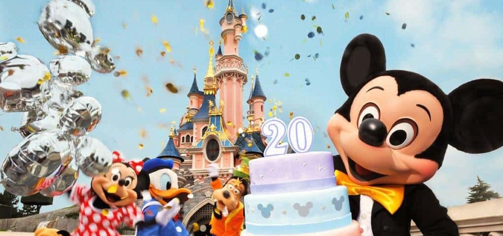 Mickey Mouse et ses amis font la fête avec un gâteau festif marqué "20" devant le château de Disneyland Paris, avec des confettis et des ballons dans les airs.