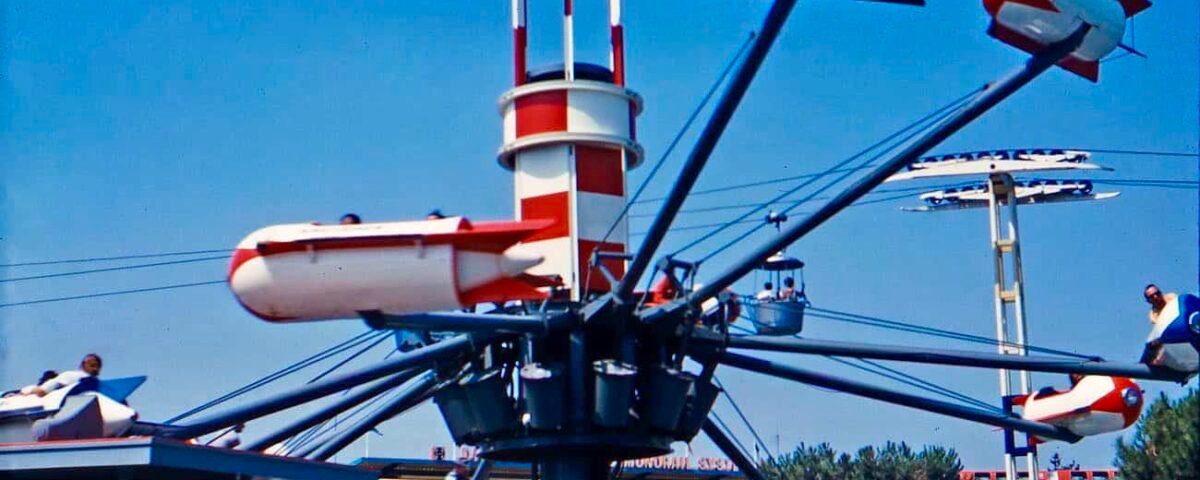 Un parc d'attractions vintage avec des modèles réduits d'avions tournant autour d'une tour centrale qui ressemble à un phare. Le trajet sous-marin d’Oswald est partiellement visible en arrière-plan sous un ciel bleu clair.
