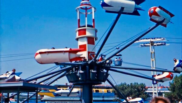 Un parc d'attractions vintage avec des modèles réduits d'avions tournant autour d'une tour centrale qui ressemble à un phare. Le trajet sous-marin d’Oswald est partiellement visible en arrière-plan sous un ciel bleu clair.