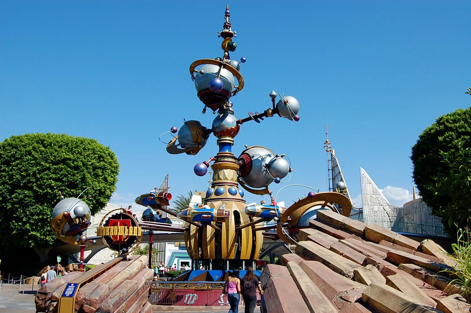 Une sculpture imaginative et futuriste ressemblant à l'Astro Orbiter dans un parc à thème, avec des couleurs vives, des éléments sphériques et des détails mécaniques sous un ciel bleu clair.