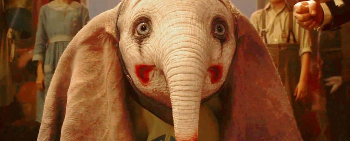 Une scène d'un film mettant en scène Dumbo, un bébé éléphant CGI aux grands yeux expressifs et aux oreilles surdimensionnées, vêtu d'une tenue colorée. Deux enfants apparaissent en arrière-plan, observant l'éléphant.