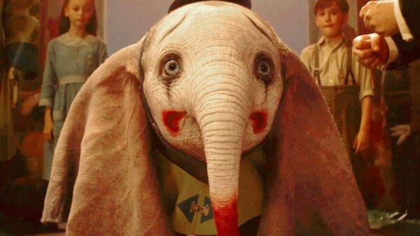 Une scène d'un film mettant en scène Dumbo, un bébé éléphant CGI aux grands yeux expressifs et aux oreilles surdimensionnées, vêtu d'une tenue colorée. Deux enfants apparaissent en arrière-plan, observant l'éléphant.