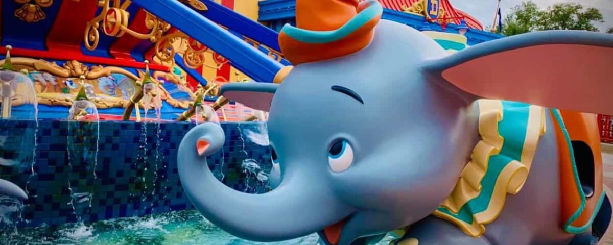 Statue colorée de Dumbo l'éléphant volant avec un chapeau orange vif, sur fond de bâtiment animé et de ciel nuageux dans un parc à thème.