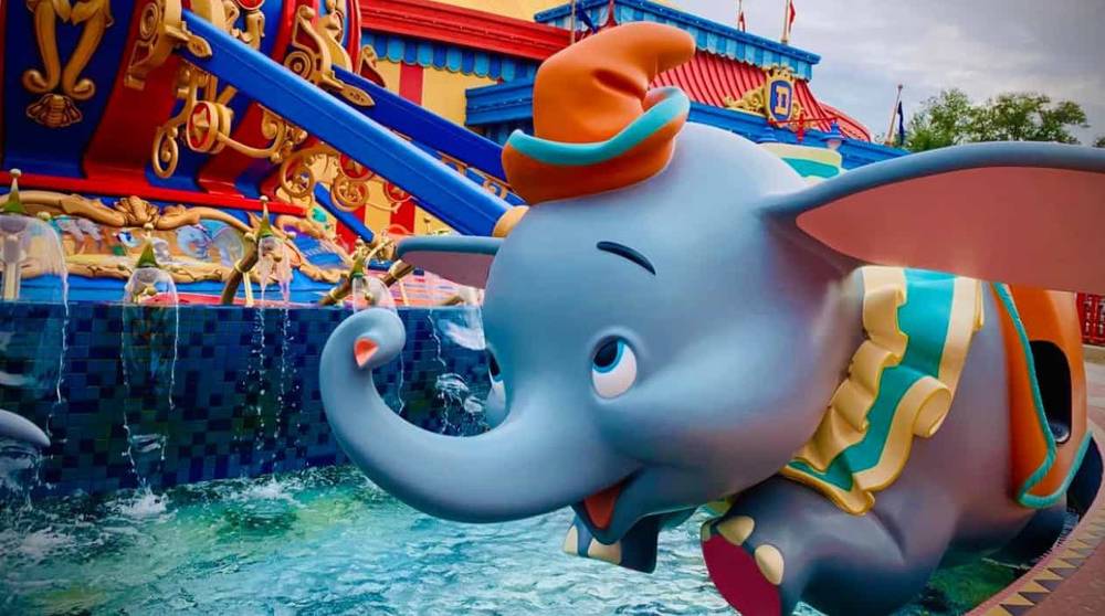 Statue colorée de Dumbo l'éléphant volant avec un chapeau orange vif, sur fond de bâtiment animé et de ciel nuageux dans un parc à thème.