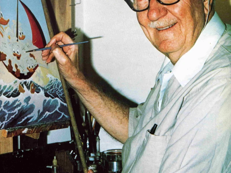 Homme âgé avec des lunettes et une casquette, souriant en peignant une scène maritime colorée sur une toile dans un studio bien éclairé, rappelant le style de Carl Barks.