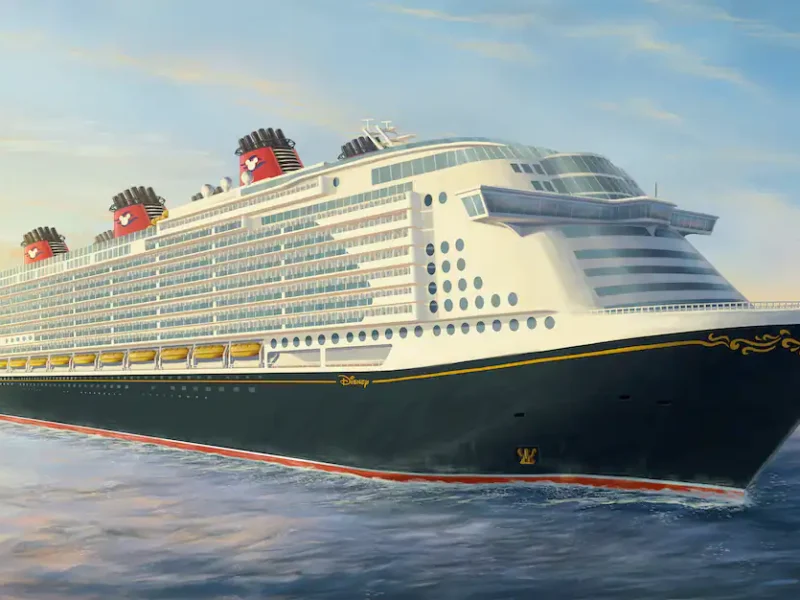 Un grand navire Disney Cruise Line naviguant sur une mer calme au coucher du soleil, avec un ciel doré et des nuages duveteux en arrière-plan.
