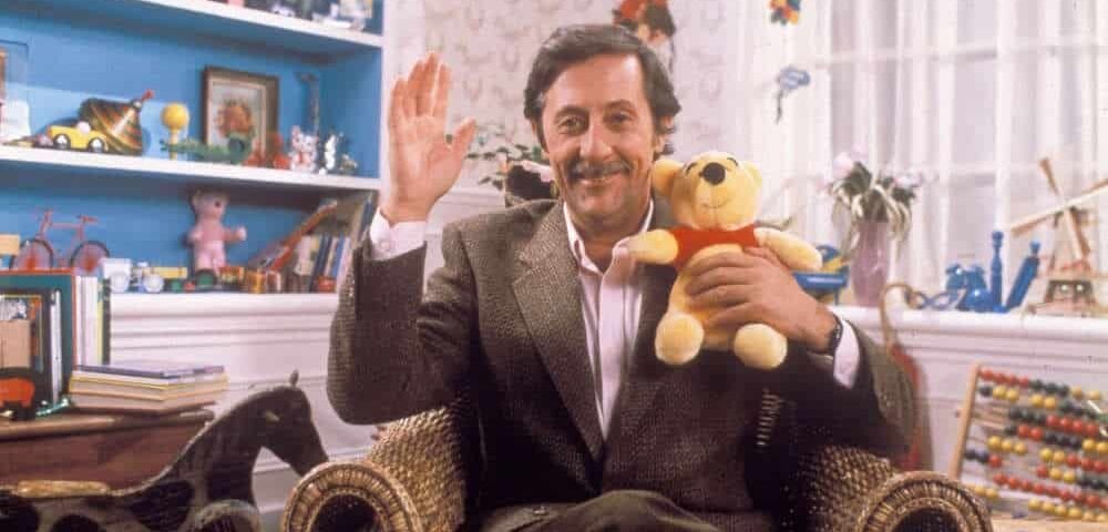 Un homme avec une barbe et une moustache, vêtu d'un costume, est assis sur une chaise en osier dans une pièce colorée remplie de jouets inspirée de Disney Channel, souriant et saluant d'une main tout en tenant