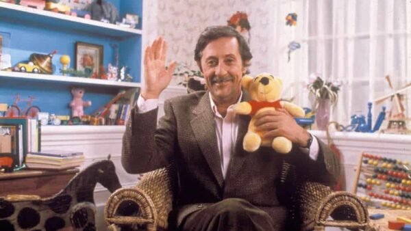 Un homme avec une barbe et une moustache, vêtu d'un costume, est assis sur une chaise en osier dans une pièce colorée remplie de jouets inspirée de Disney Channel, souriant et saluant d'une main tout en tenant