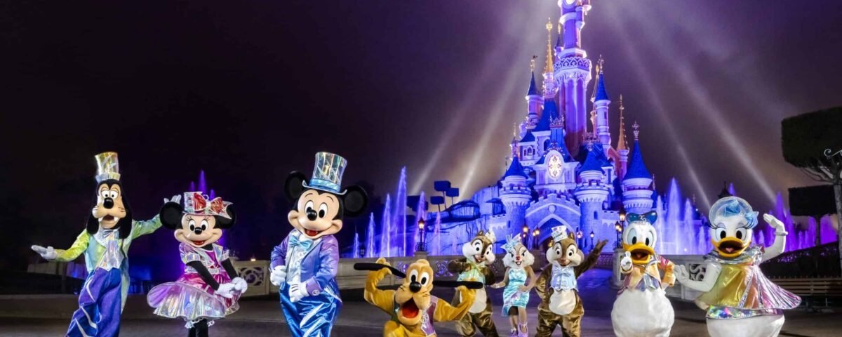 Mickey Mouse et ses amis vêtus de costumes colorés devant le château de Cendrillon à Disneyland Paris, éclairé par des projecteurs sur fond de brume lors de leur célébration d'anniversaire.
