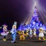Mickey Mouse et ses amis vêtus de costumes colorés devant le château de Cendrillon à Disneyland Paris, éclairé par des projecteurs sur fond de brume lors de leur célébration d'anniversaire.